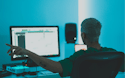 Instant online audio mastering imusician hero image Computer monitor, keyboard, and person's back in dark office setting with blue lighting.