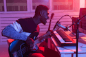 Entender YouTube Content ID Un músico canta apasionadamente en un micrófono mientras toca la guitarra. Está sentado en un escritorio con un teclado, en una habitación iluminada con luces rojas y rosas, creando un ambiente cálido y creativo.